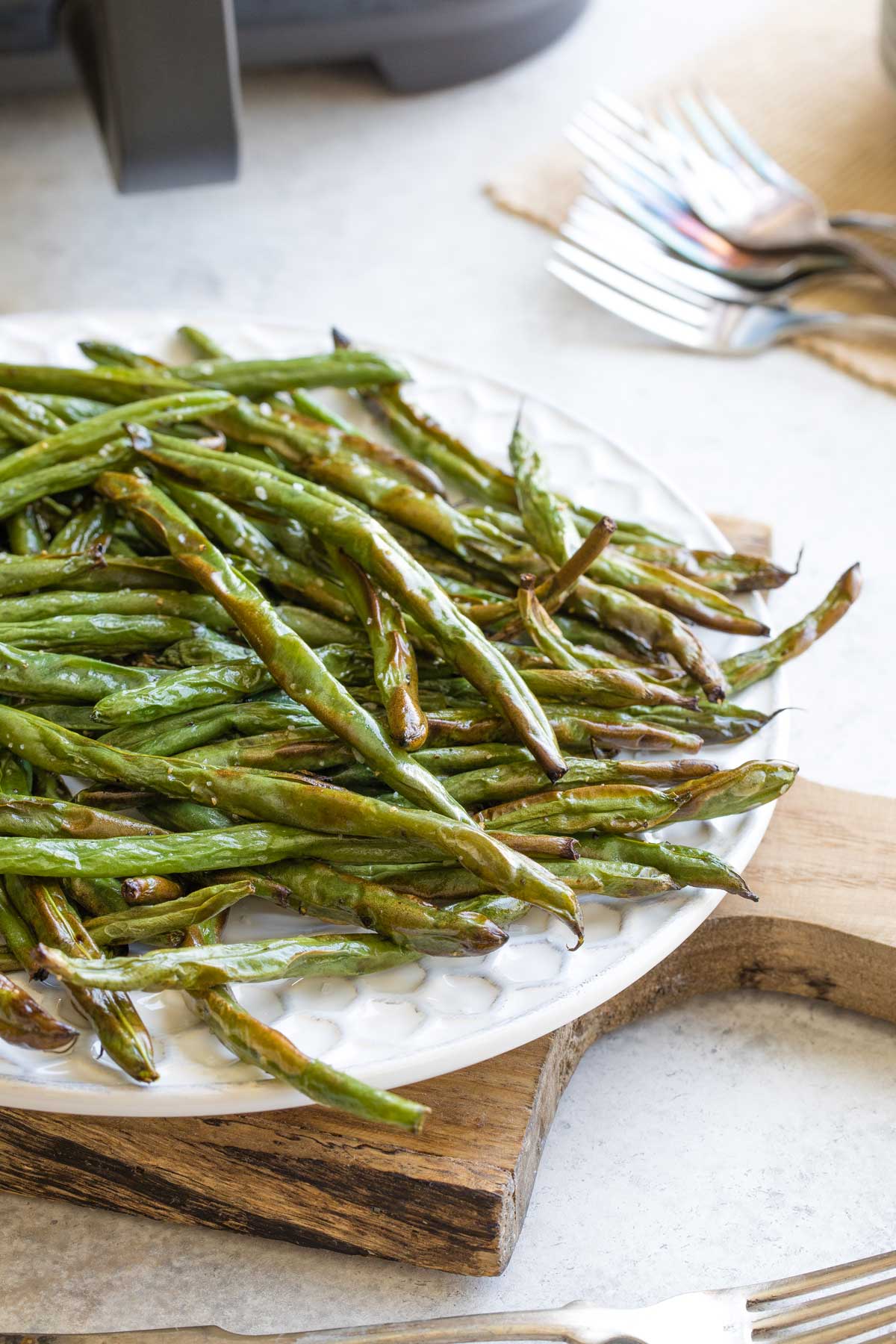 Side view of right half of platter heaped with air fried green beans, with air fryer and extra forks in background.