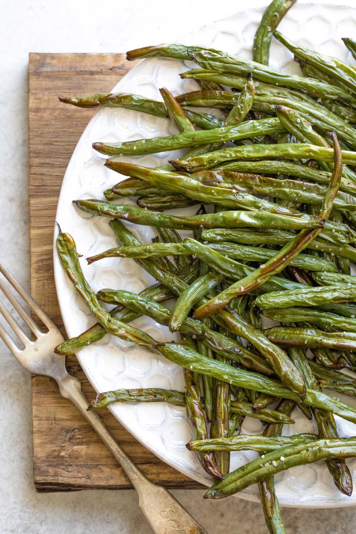 Overhead of left side of serving platter full of air fried green beans, with antique fork alongside on wooden board.