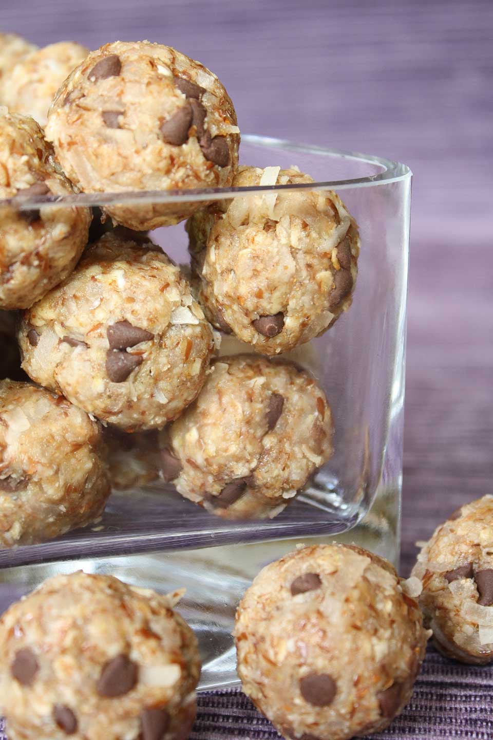 A dozen or so finished energy bites in a clear glass triangular dish, with 3 laying in front - all on purple cloth backdrop.