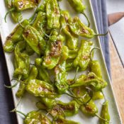 Overhead of a platter of shishito peppers laying on gray cloth on a wooden tray.