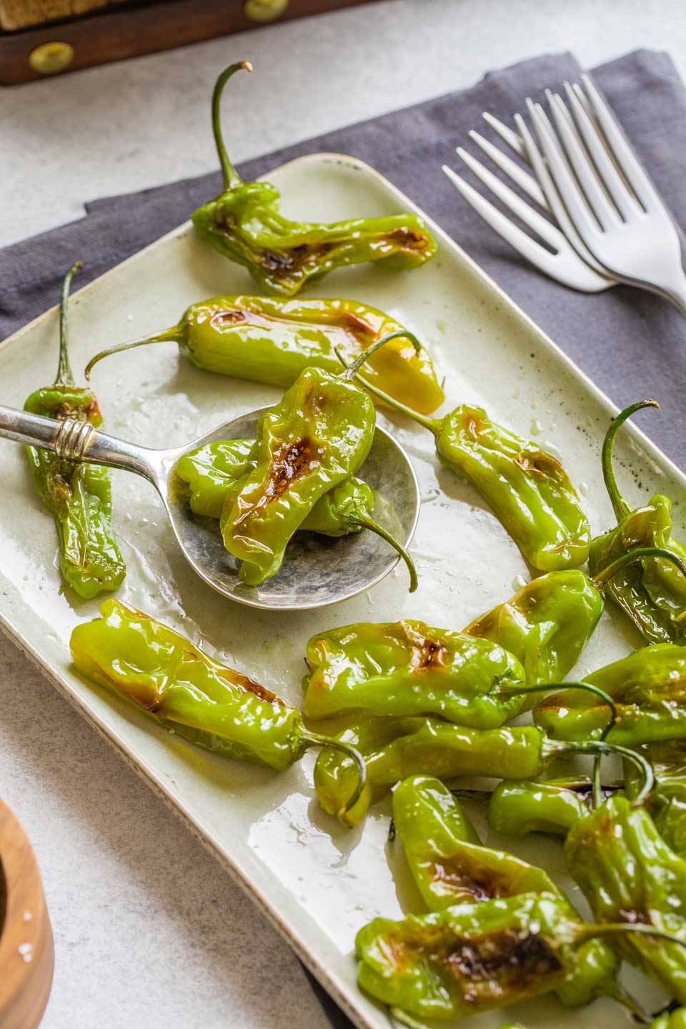 Two blistered shishitos on a serving spoon, resting on serving platter with other peppers nearby.