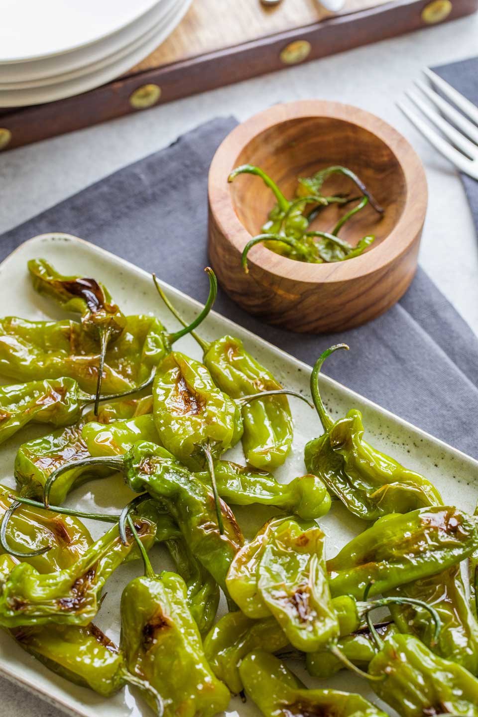 Seared peppers on serving plate with wooden bowl for stems nearby.