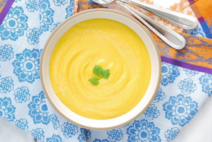 Flatlay of tan-rimmed bowl of soup garnished with fresh herb, on blue, purple and orange patterned cloth.