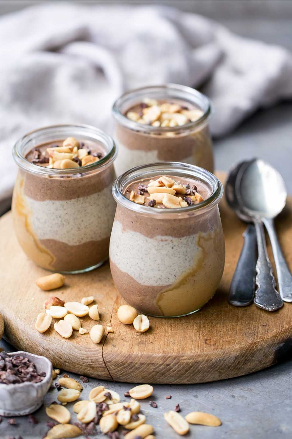 3 jars of swirled chia seed pudding on a round wooden board with spoons and peanuts nearby.