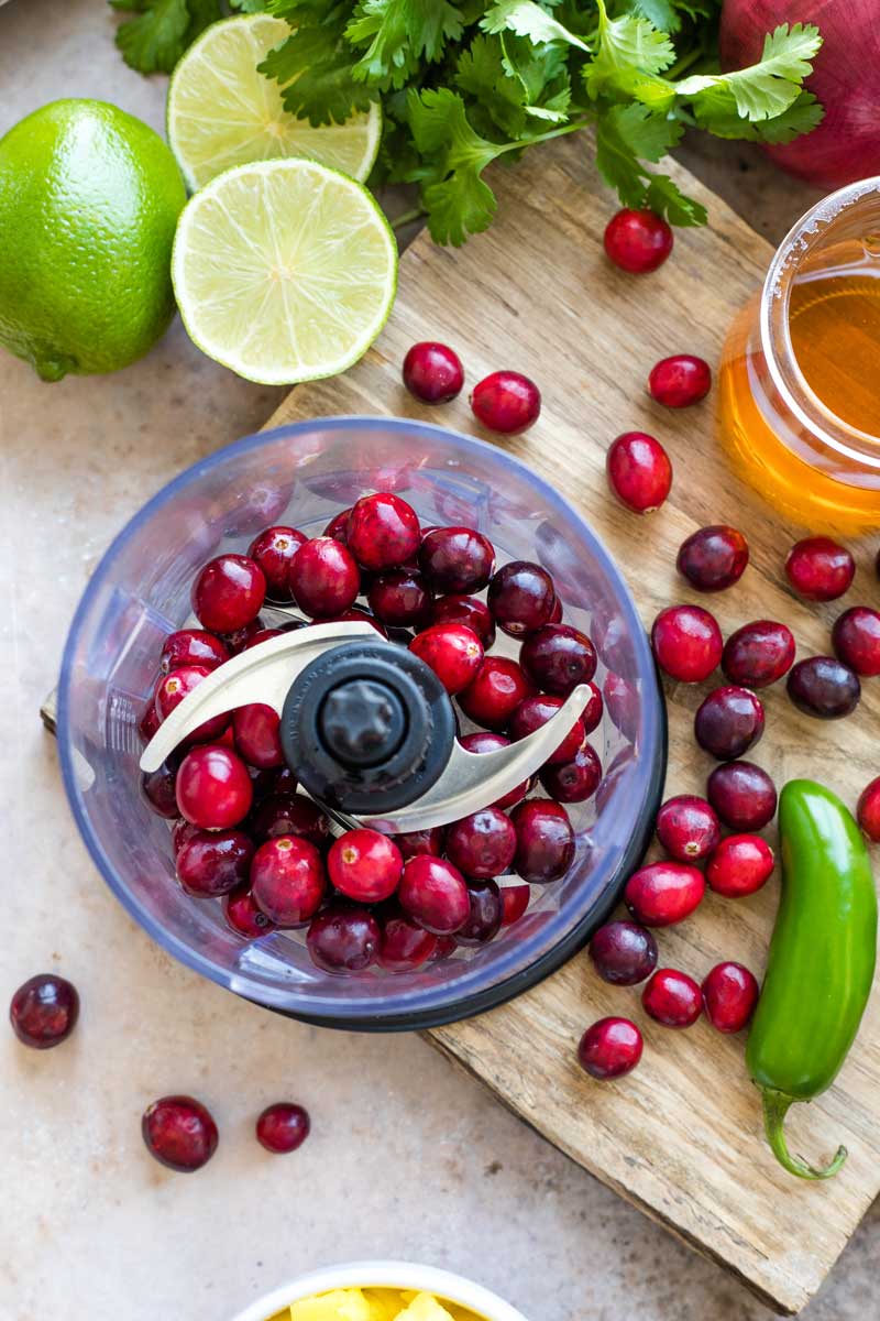 Overhead of mini food processor with whole cranberries inside, amidst other ingredients on cutting board.