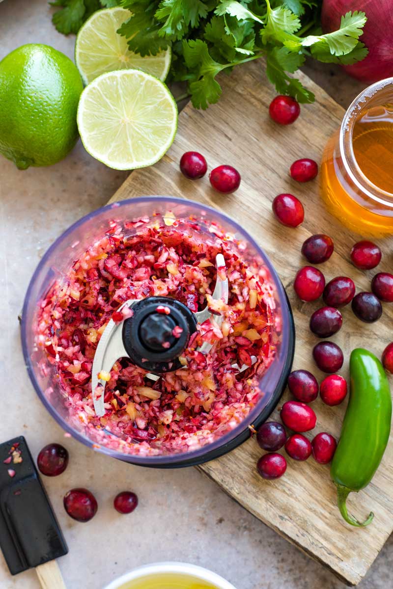 Overhead of processor with completely chopped cranberry and pineapple mixture, surrounded by other ingredients on cutting board.