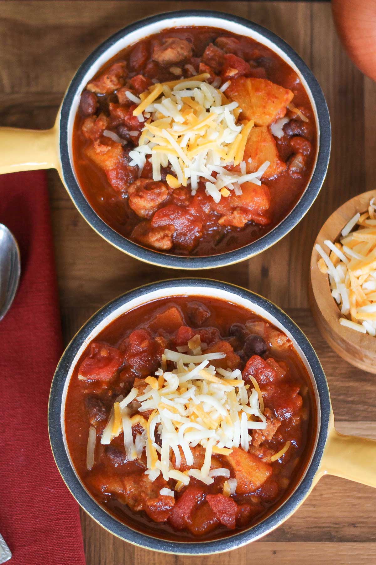 Overhead of two yellow enameled crocks full of chili,. topped with shredded cheese with a little wooden bowl of cheese, a spoon and burgundy napkin alongside.