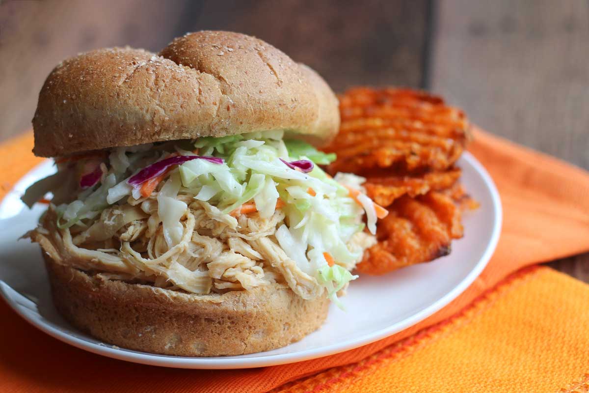 Side closeup of one BBQ chicken sandwich topped with coleslaw on a white plate with a few sweet potato waffle fries alongside.