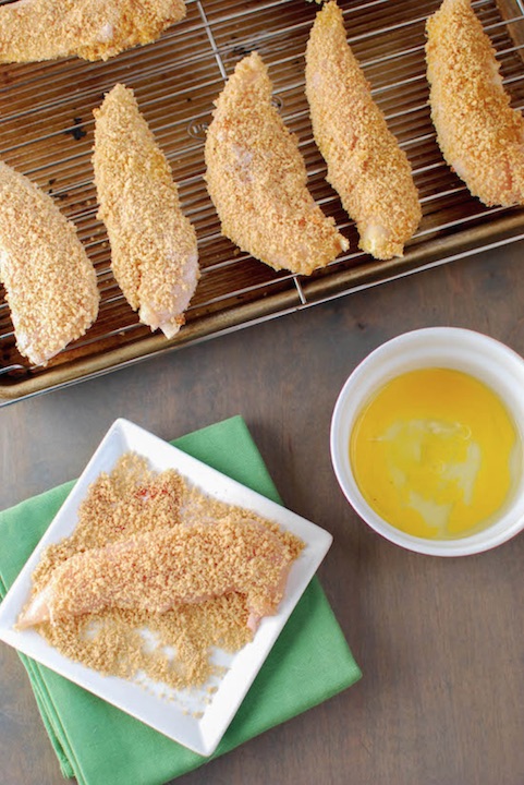 Flatlay of one tender being dredged in breadcrumbs, with blow of egg an baking sheet of raw, breaded tenders surrounding.