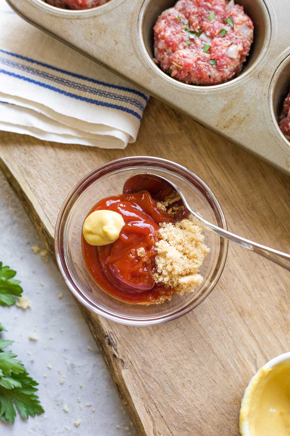 Small bowl with glaze ingredients ready to mix, next to muffin tin on cutting board.