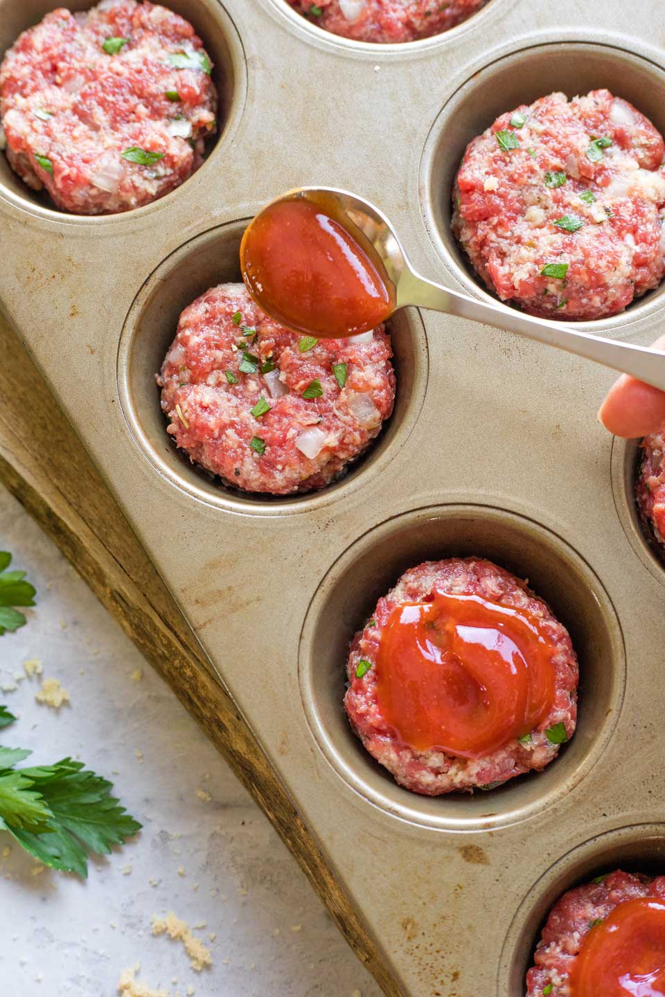 Spoon dropping glaze on top of mini meatloaf in muffin tin pan before baking.