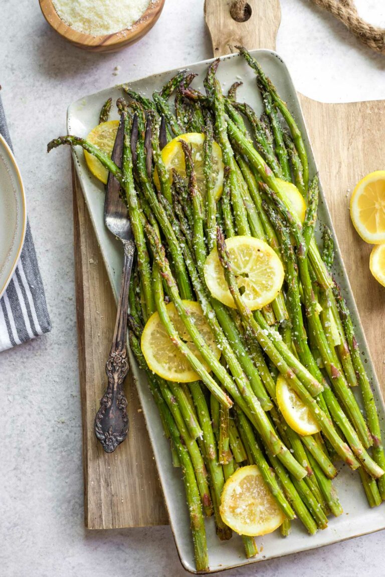 Overhead of grilled asparagus on rectangular platter with lemons and antique fork tucked in; parmesan nearby.