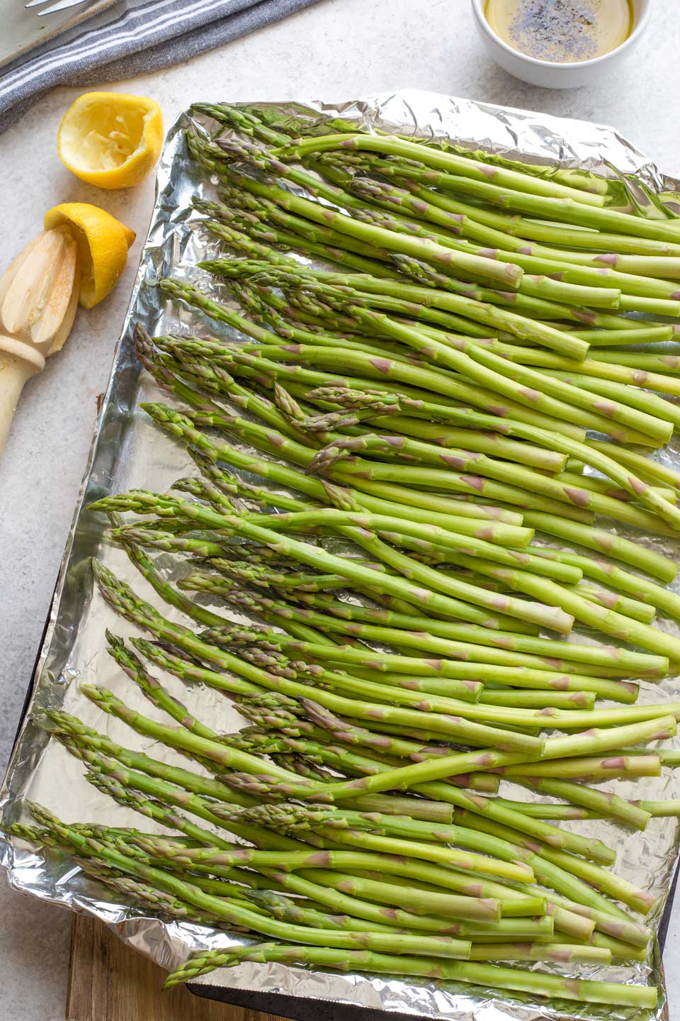 Overhead of raw asparagus spread out on heavy duty foil on grill pan; bowl of marinade in background.