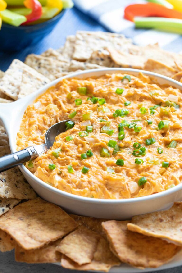 Healthy Buffalo Chicken Dip recipe presented for a party, with spoon in, chips all around, and vegetable strips for dipping in the background.