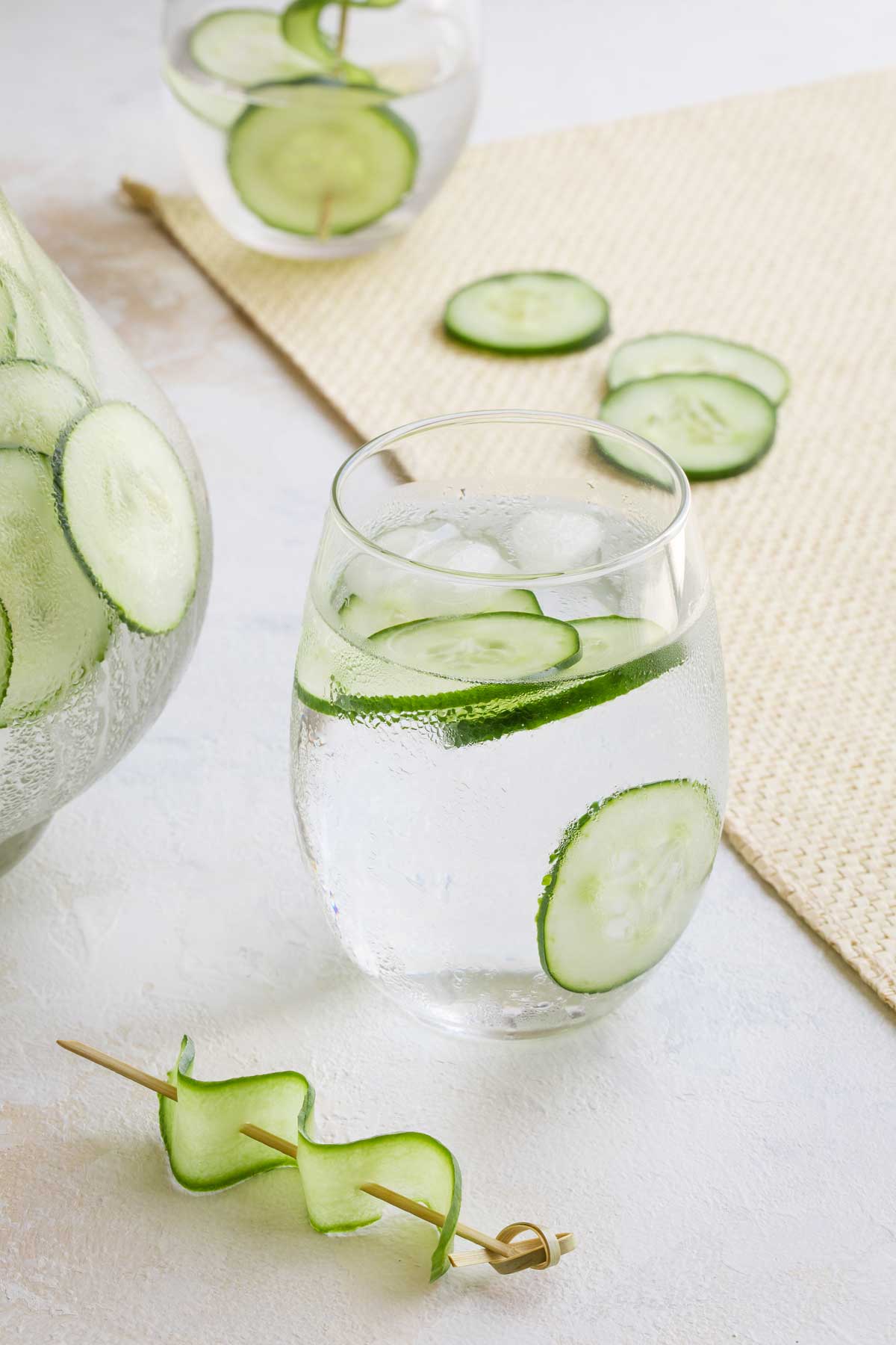 Side shot of one filled glass, with another glass, part of a pitcher, extra cucumber slices and garnish surrounding.
