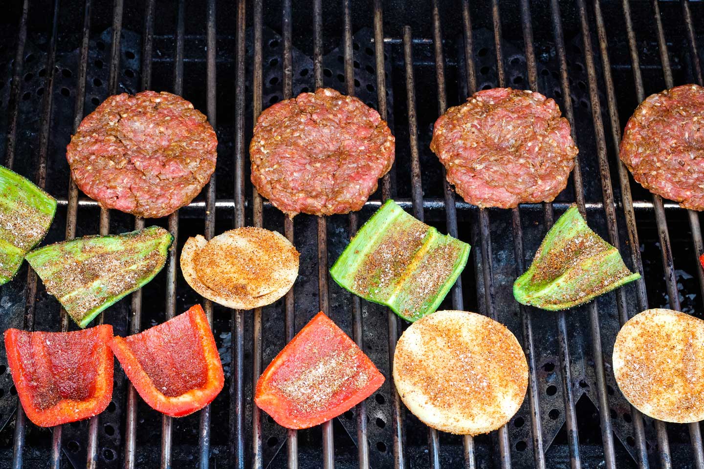 four hamburgers plus seasoned peppers and onions, cooking on outdoor grill