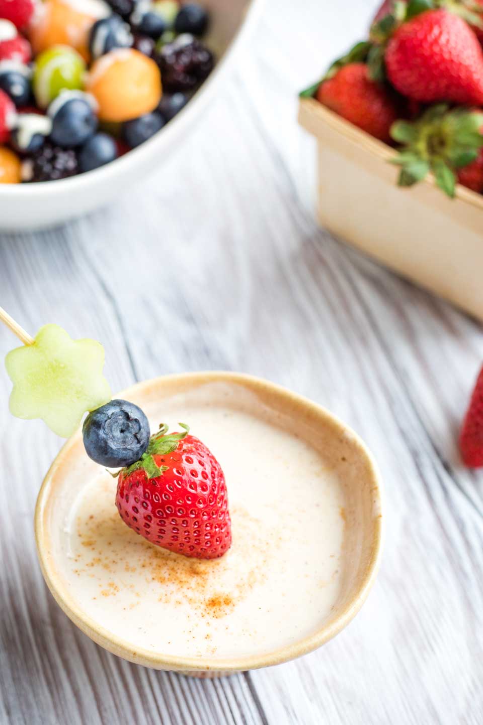 little bowl of dip with a fruit skewer containing a strawberry, a blueberry and a honeydew flower dipping into it, with other fruit in the background