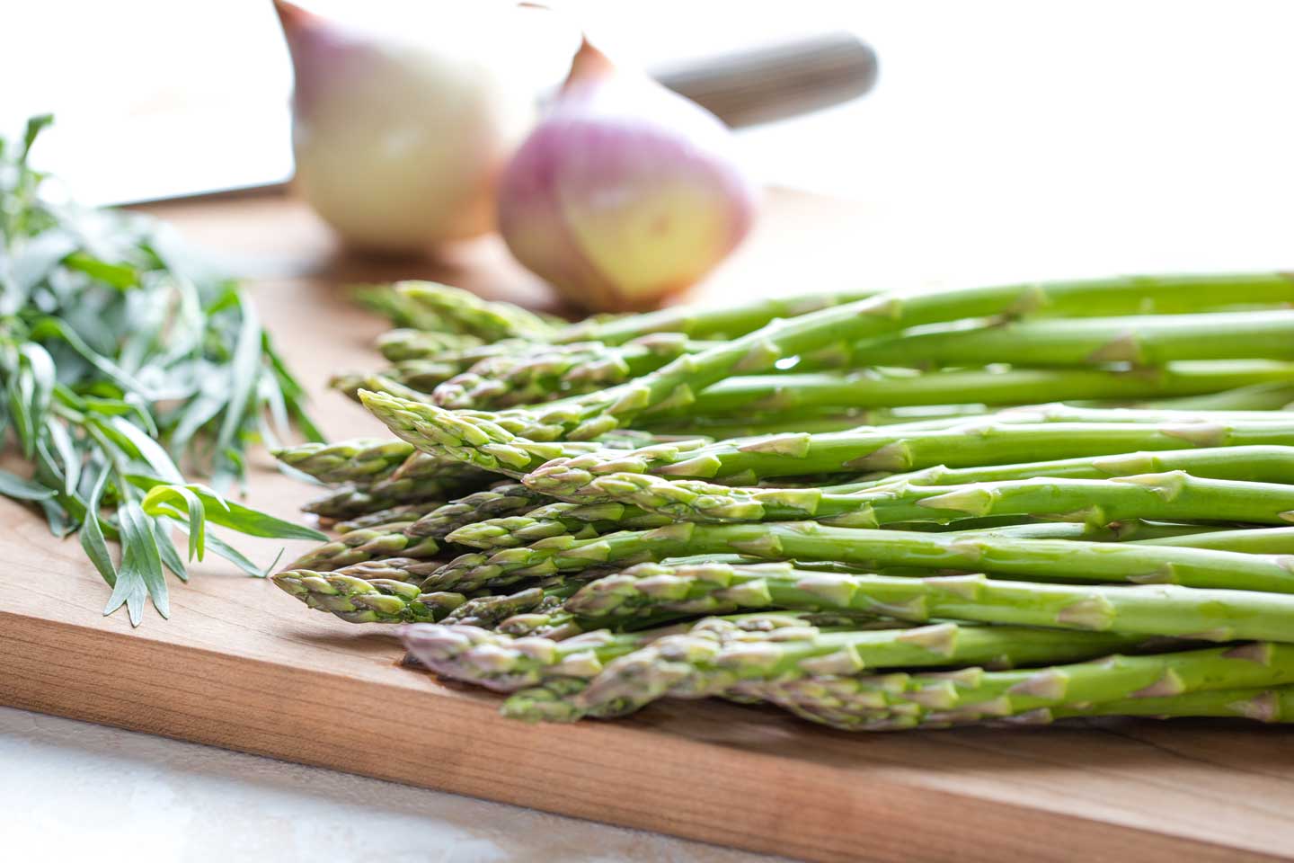 Closeup side view of fresh asparagus spears with herbs, a knife and other vegetables in the background.