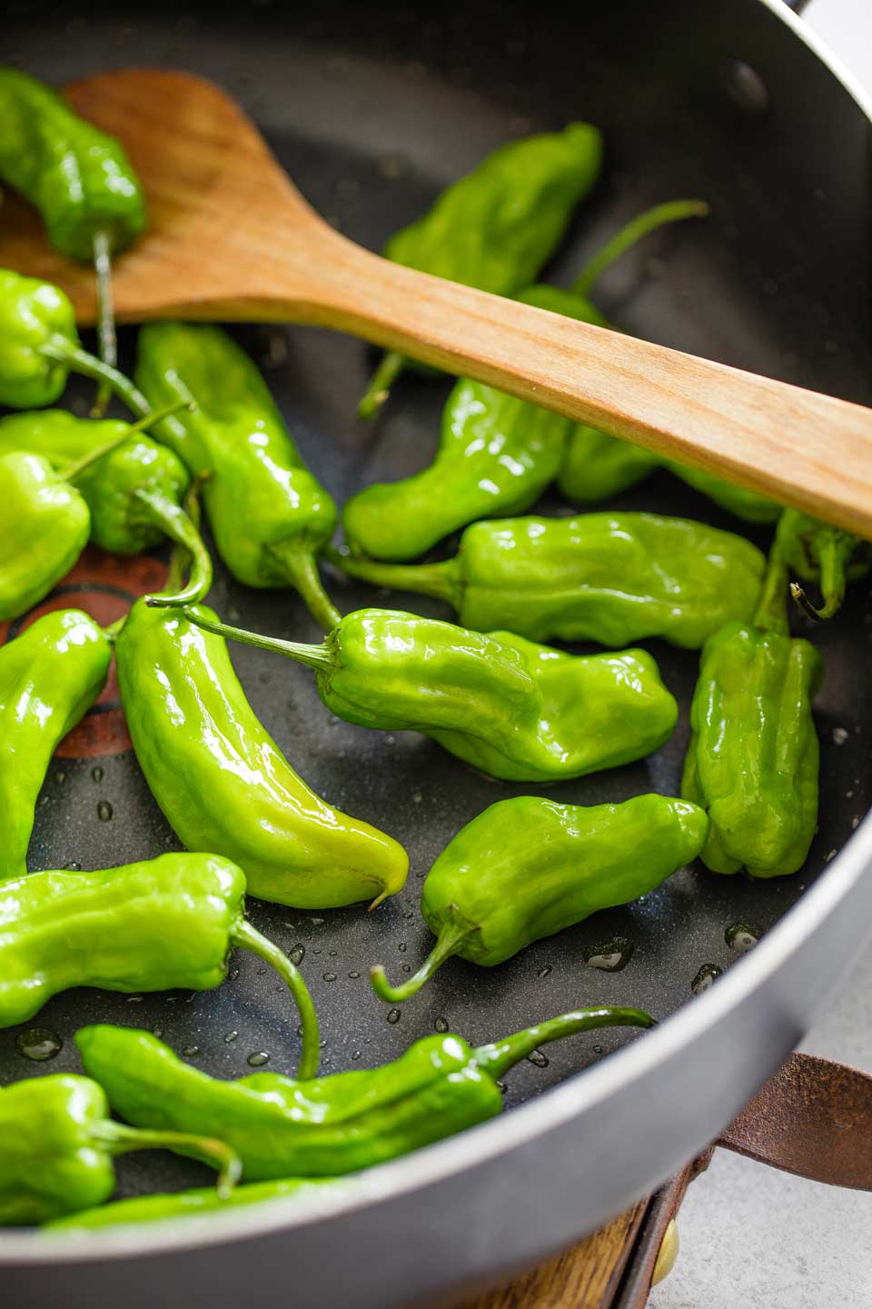 Closeup of part of a nonstick skillet where a wooden spoon is stirring shishitos as they begin to cook.