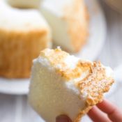 Closeup of one slice of cake being held up by a hand in the foreground, with the rest of the uncut cake on a white platter in the background.