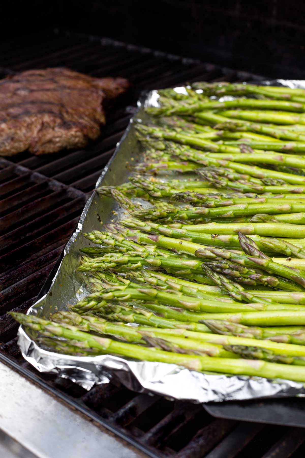 Side view of tops of asparagus in pan on grill with steak cooking behind.