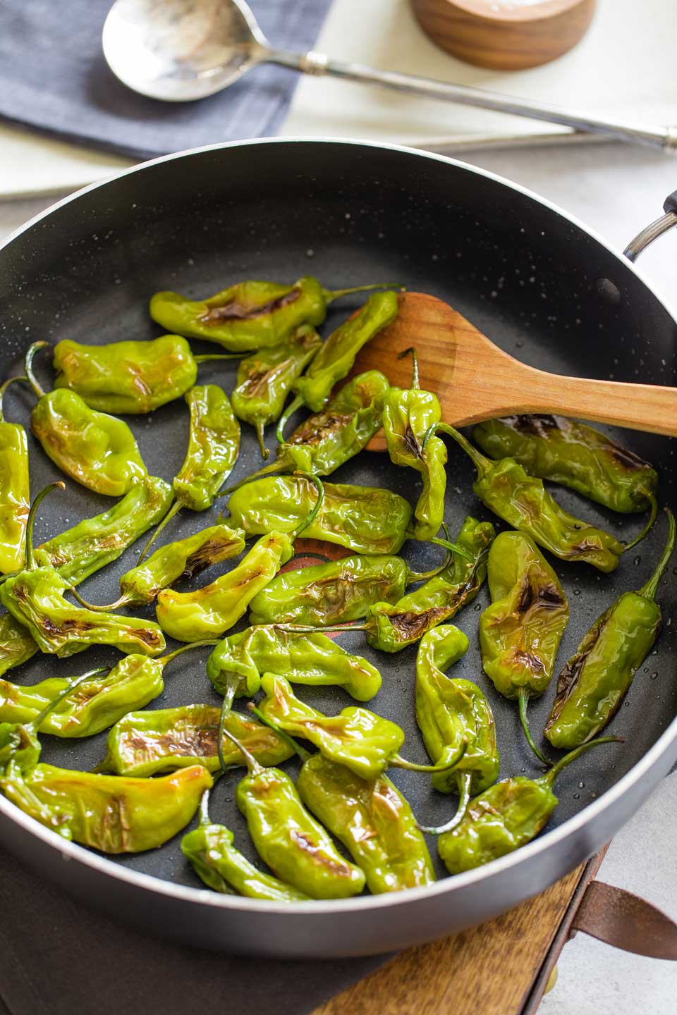 Overhead of wooden spoon stirring fully cooked, blistered shishitos in pan.