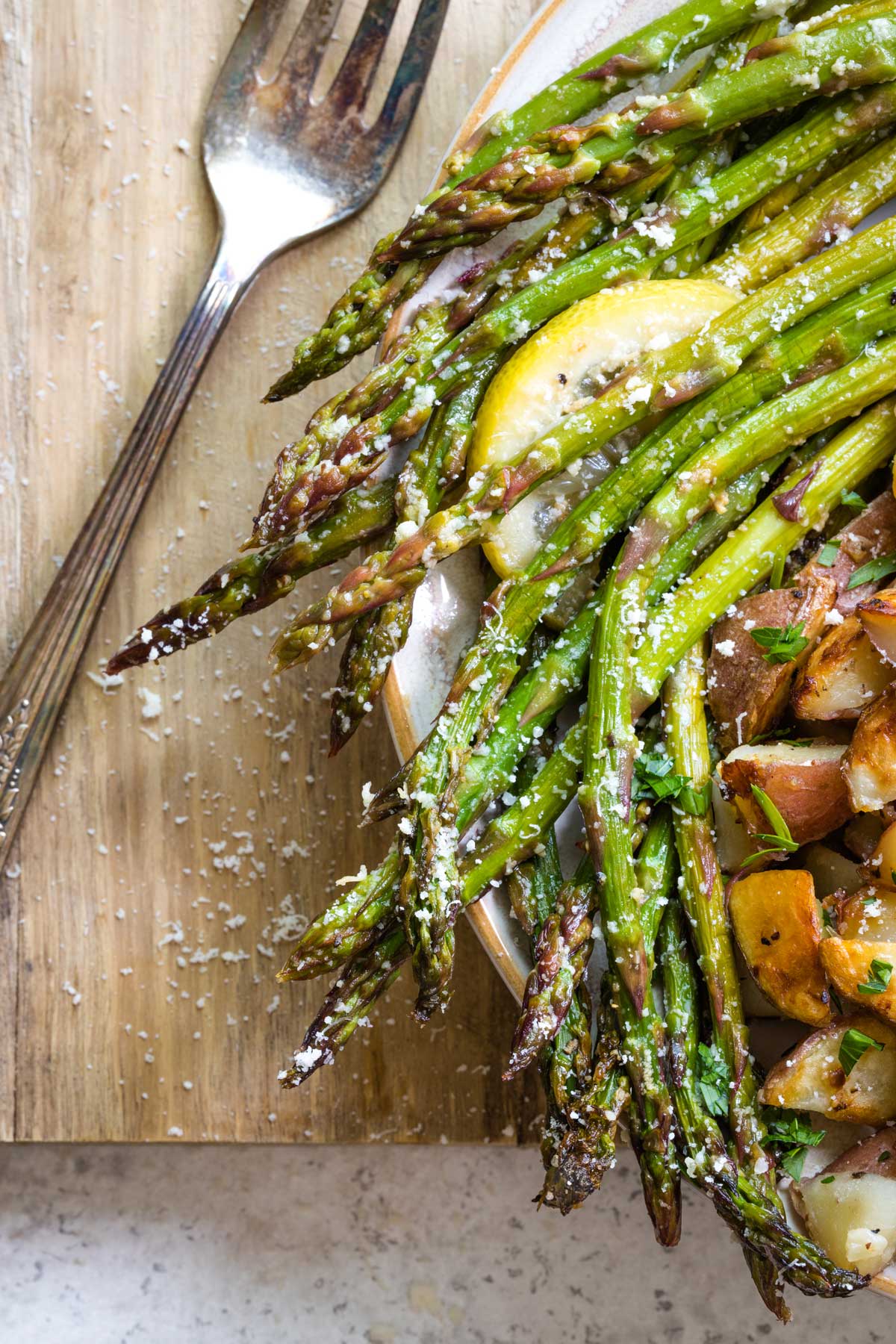 Closeup of Asparagus sprinkled with parmesan at edge of dinner plate with potatoes; fork nearby.