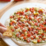 Fingers dipping pita chip into pesto blob at edge of finished appetizer on white plate on wooden board; more crackers and mini peppers in background.