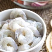 Closeup of a white bowl filled with raw, thawed shrimp, with other ingredients for a recipe at the edges of the photo.