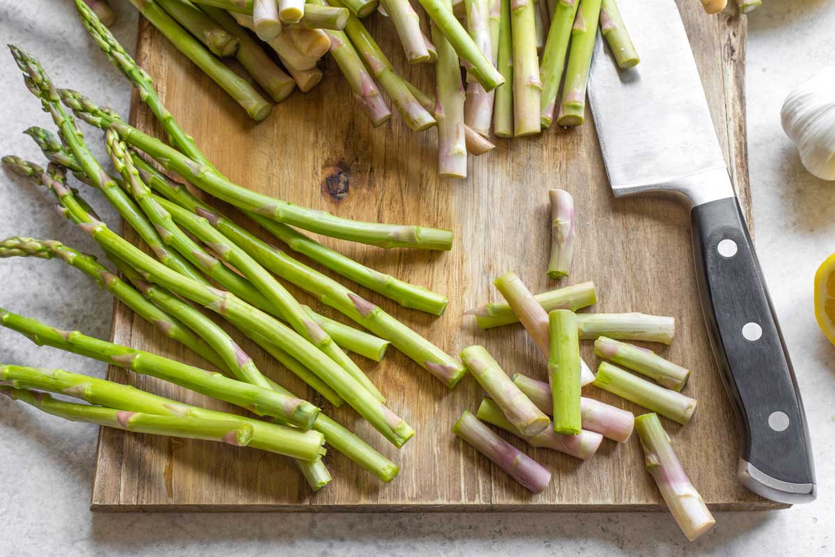 Raw asparagus spears on wood cutting board, some with ends trimmed off by knife.