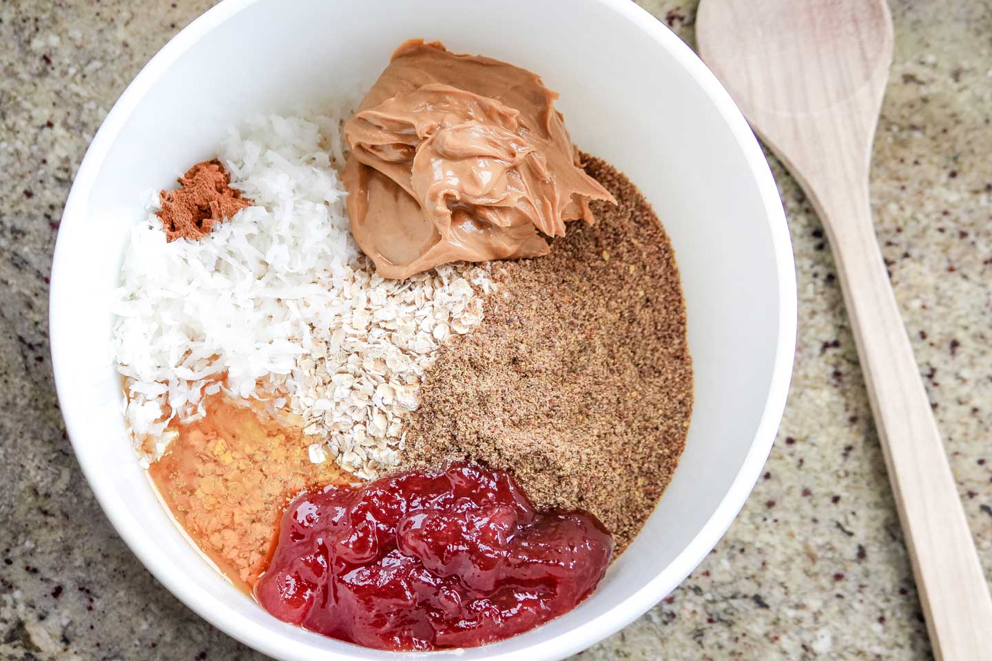 All the Energy Ball ingredients in separate little areas in a white bowl, with a wooden spoon alongside.