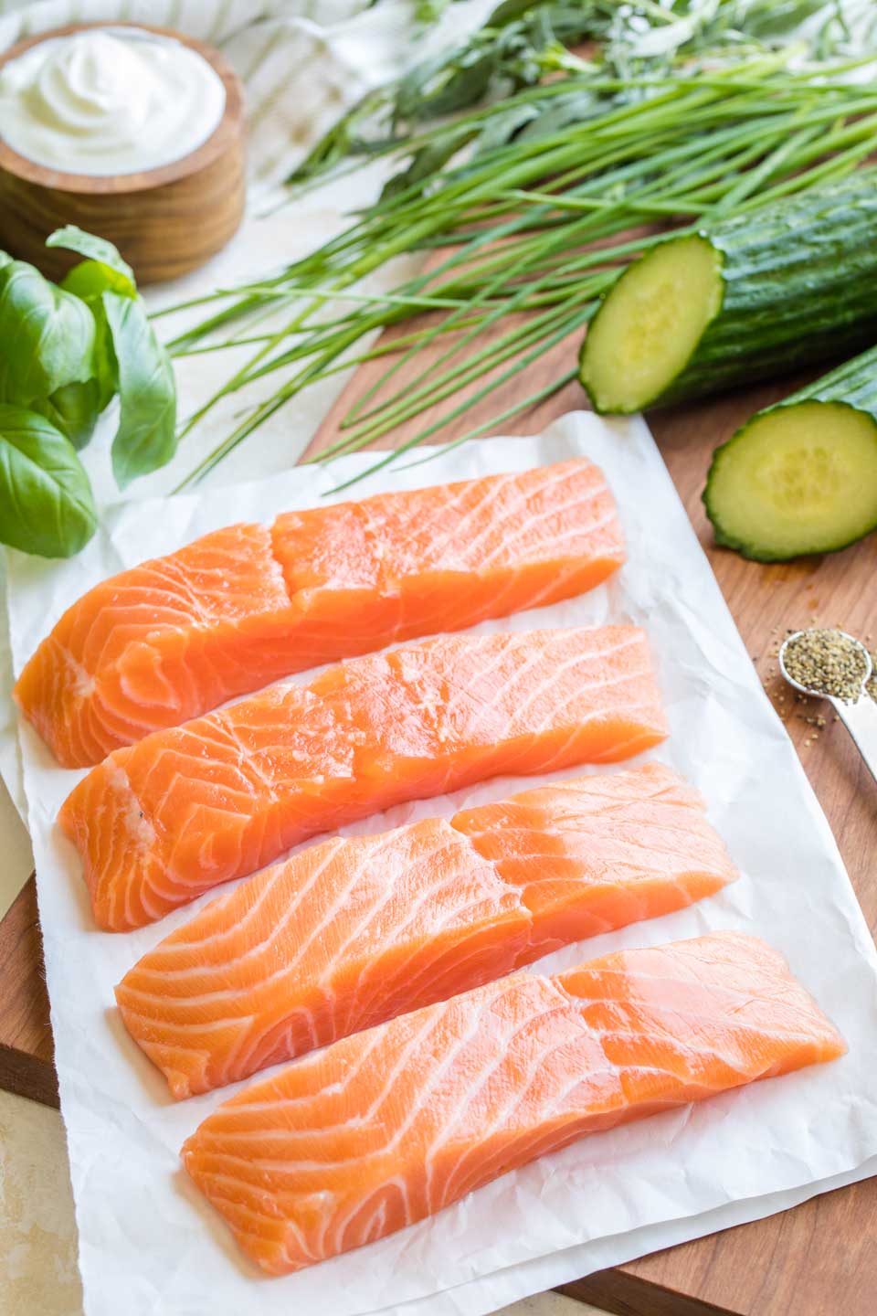 Four raw salmon fillets on white butcher paper, laying on a cutting board and surrounded by ingredients for the tzatziki sauce.