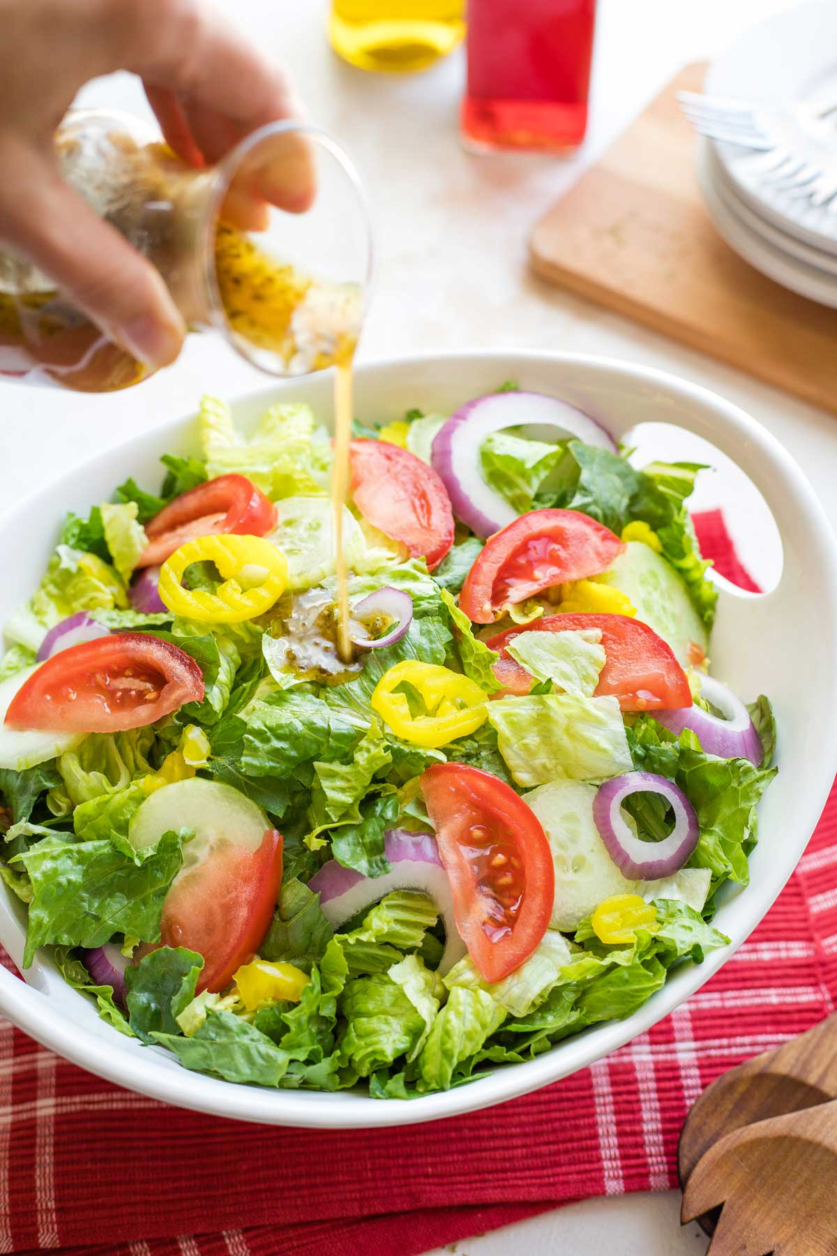 Hand pouring dressing onto bowl of green Italian salad sitting on red cloth with servers, plates, oil and vinegar nearby.