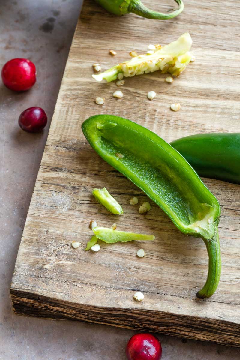 Halved jalapeno on cutting board with seeds and ribs removed and divided, and a couple cranberry nearby.