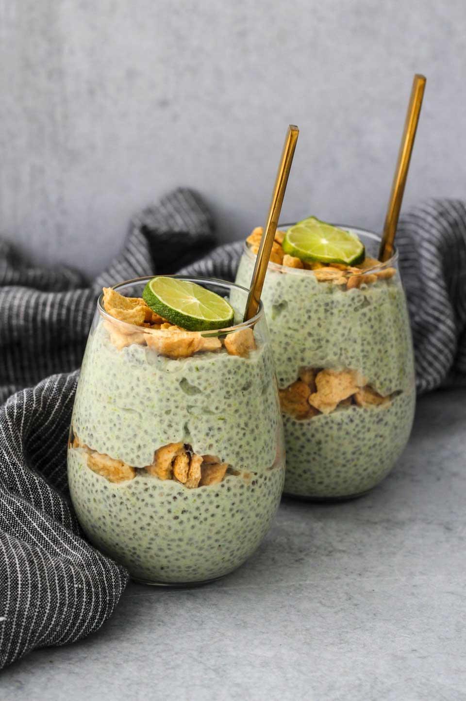 Two stemless glasses showing alternating layers of the chia pudding and broken graham crackers, with lime slices on top and gold spoons.