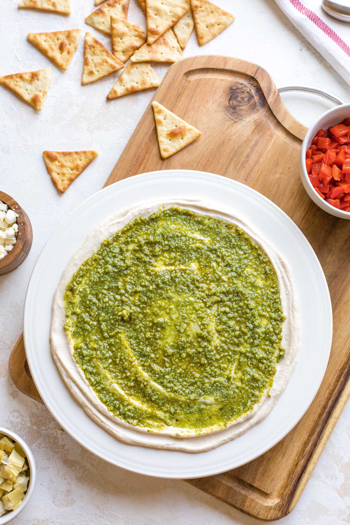 Overhead of pesto layer spread on top of cream cheese layer with other recipe ingredients nearby.