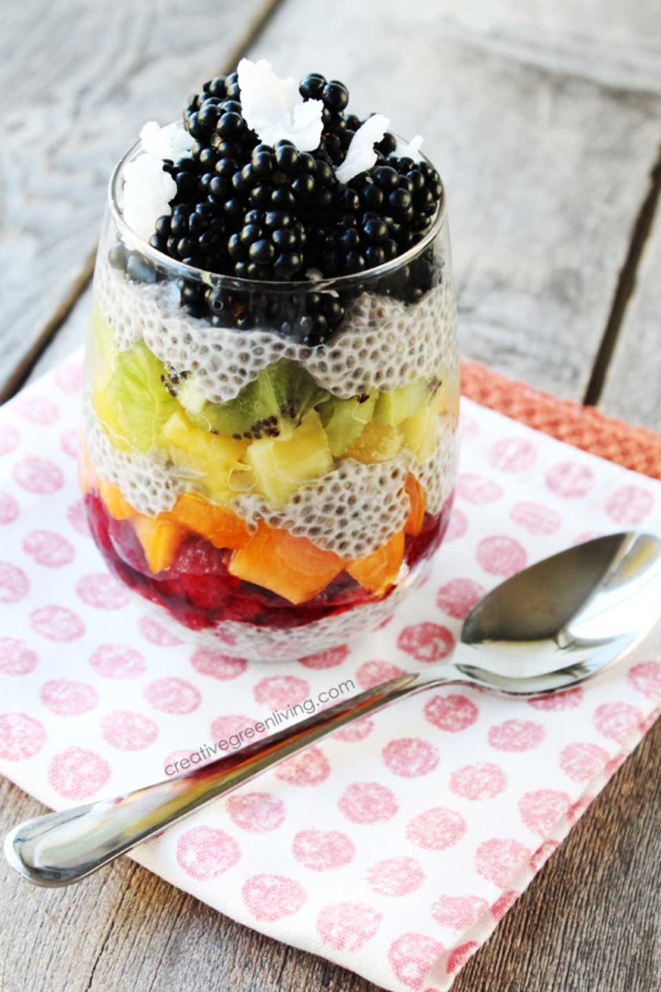 Glass with layers of chia pudding and rainbow-colored fruits, sitting on a pink polka-dot napkin next to a spoon.