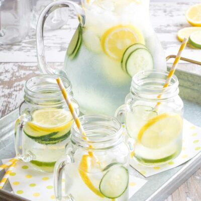 Metal tray with pitcher of infused water and three mason jar glasses, striped straws and cucumber and lemon slices.