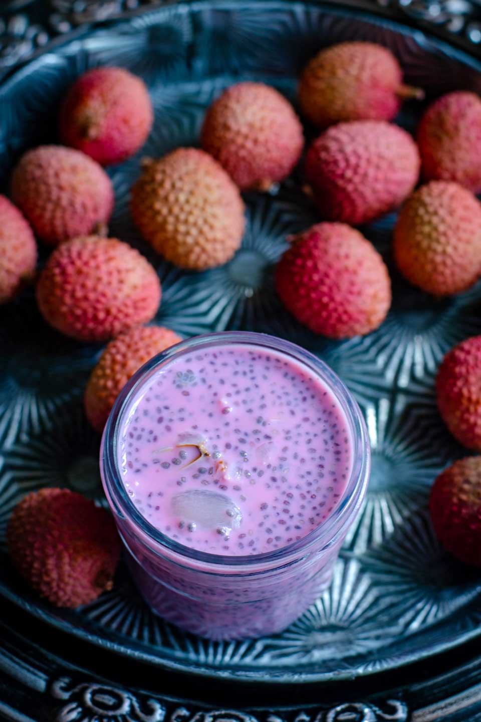 Pink chia pudding with peeled lychee floating in it, on a decorative metal tray surrounded by lychee fruit.