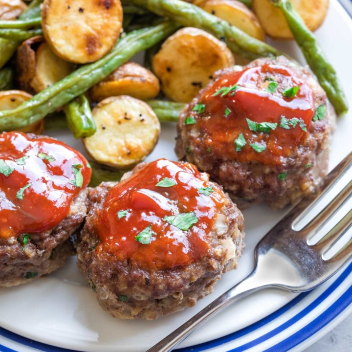 Close shot of one mini meatloaf, with two more and veggies, all plated with fork and ready to eat