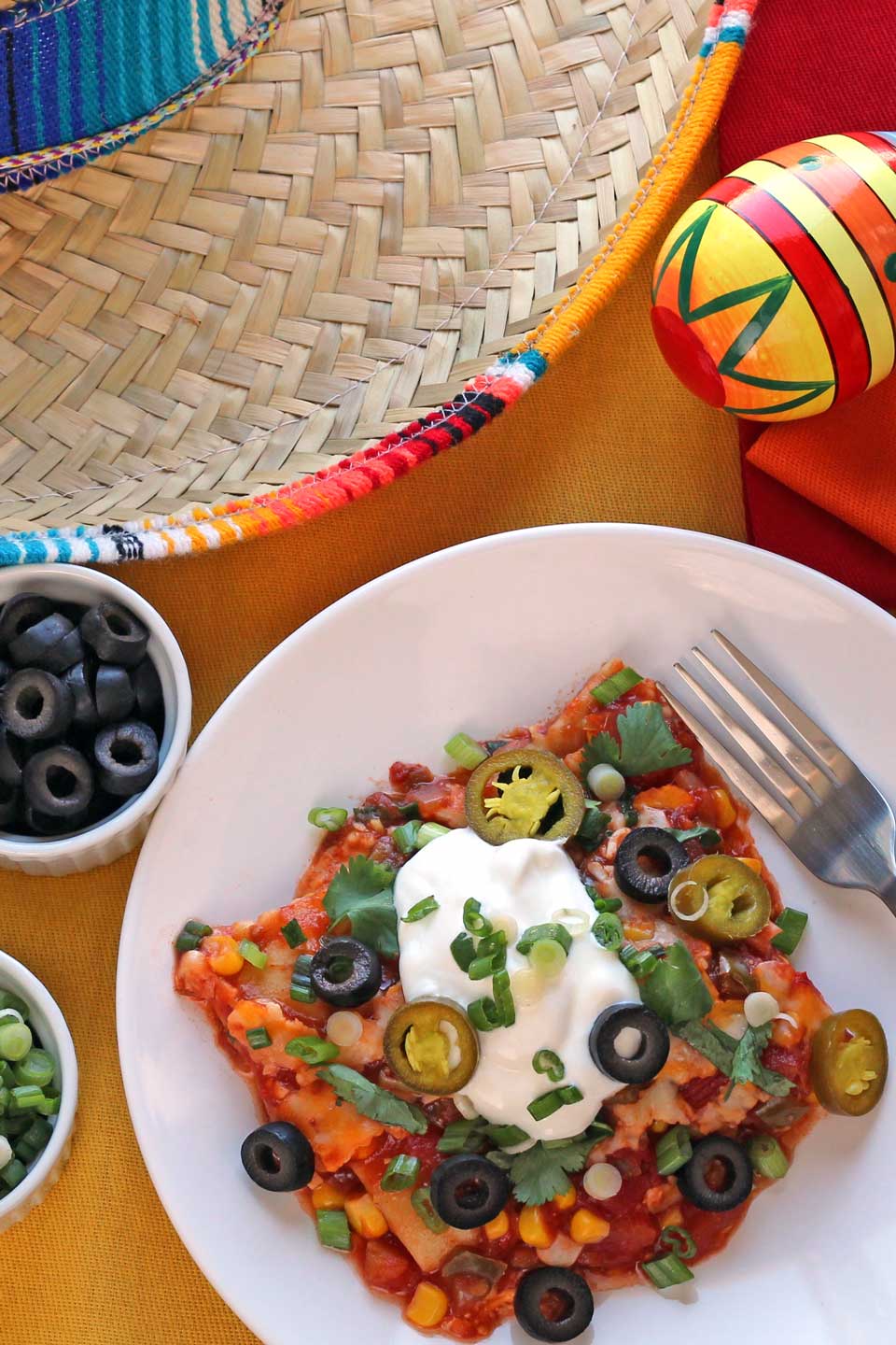 Overhead photo of one slice on this Mexican lasagna on a little white plate, surrounded by toppings and by a sombrero and maraca.