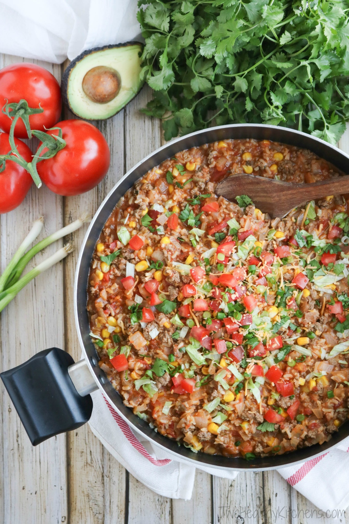 Flatlay of finished Mexican rice still in pot with toppings and wooden spoon; whole veggies and cilantro bunch nearby.