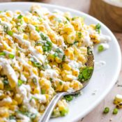 Closeup of about half the serving platter of Corn Salad, with a serving spoon dipped into the side, and squeezed lime rinds, extra cheese and cilantro leaves in the background.