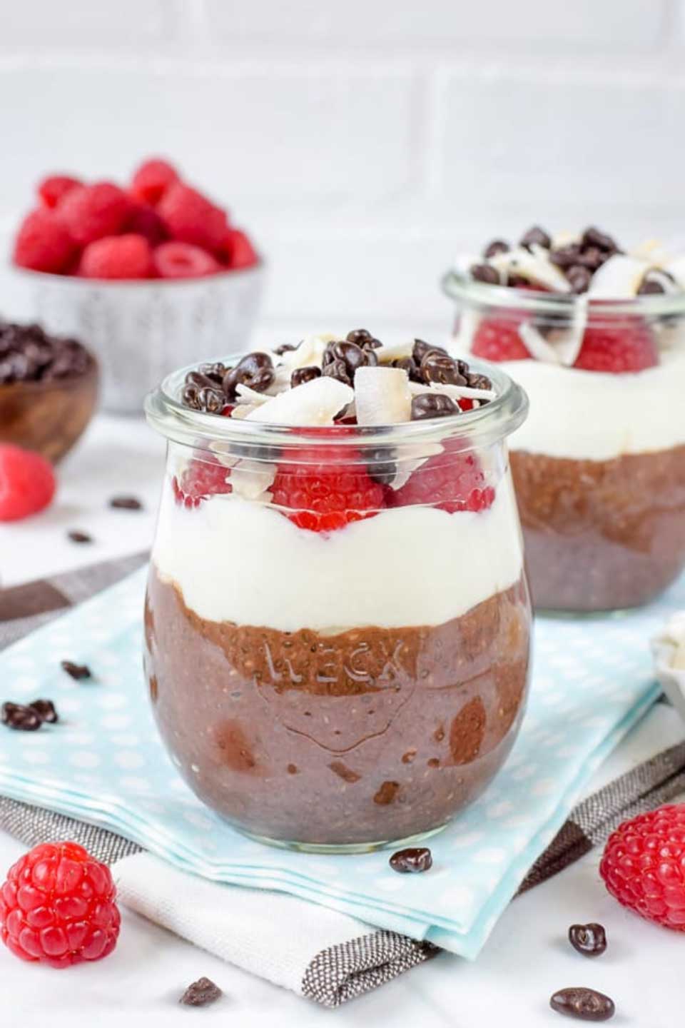 2 parfaits in Weck jars, laying on blue and black checked towels with raspberries scattered around.