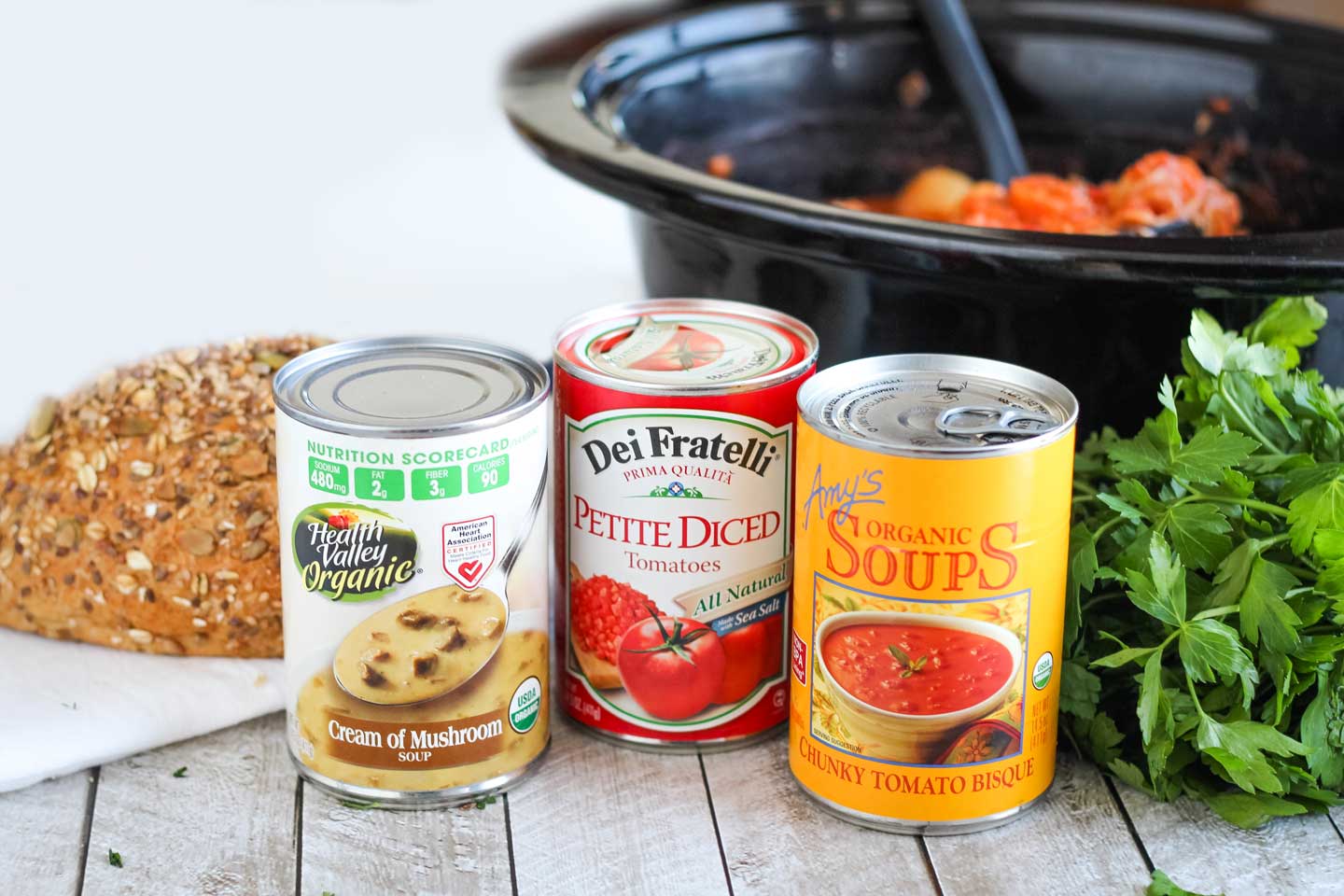 canned ingredients for a slow cooker chicken stew, with crock-pot insert and a loaf of whole grain bread in the background