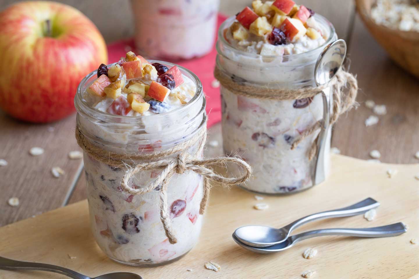 Two jars of Oats on a wooden cutting board with spoons and an uncut apple nearby.