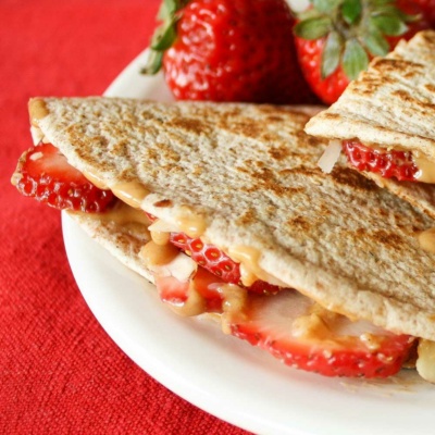 Closeup of one stuffed tortilla wedge with drippy peanut butter and whole strawberries on white plate and red fabric.