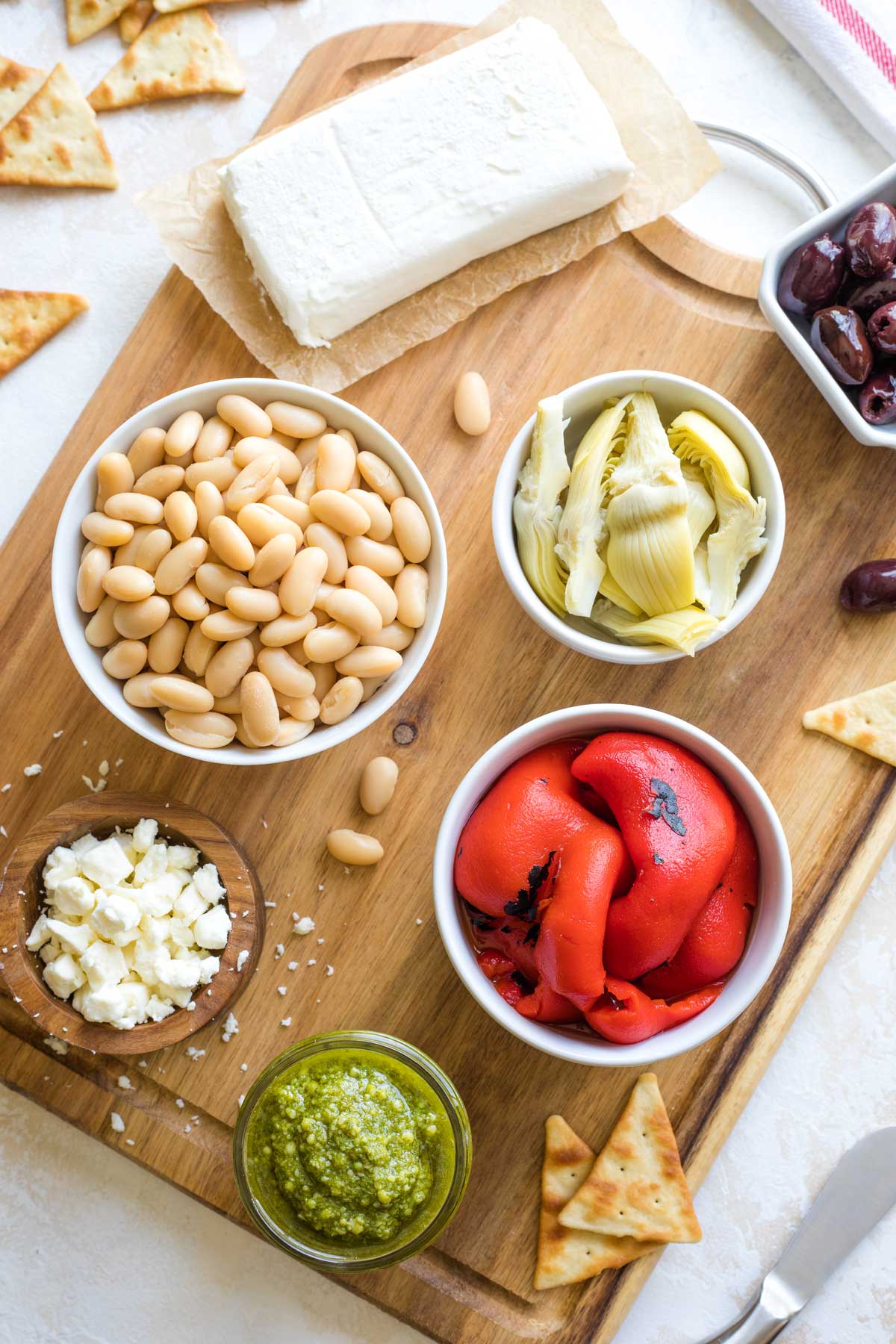 Overhead of recipe ingredients on wooden cutting board - pesto in little jar, veggies in white bowl, cream cheese on parchment paper, and appetizer spreader at corner.