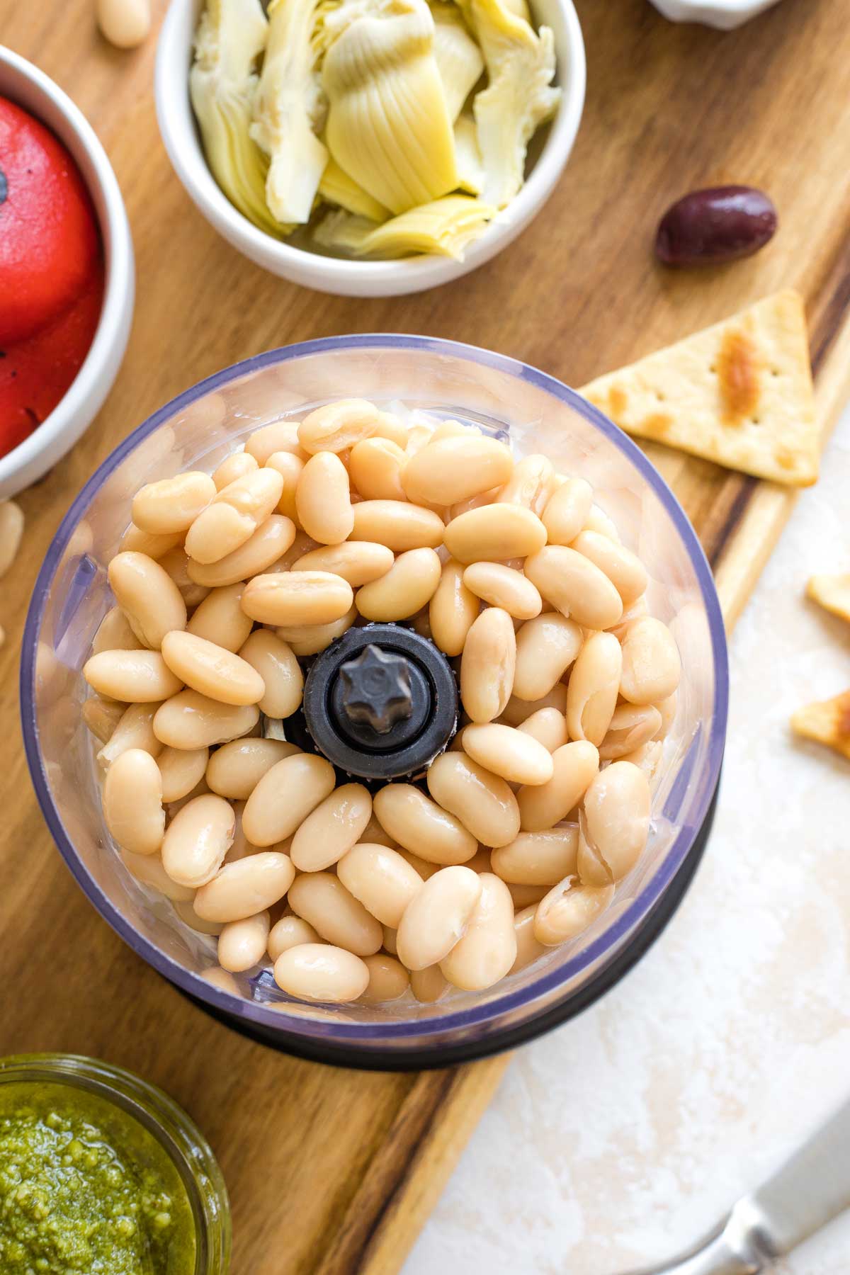 Overhead of cannellini beans on top of cream cheese in mini food processor with other ingredients surrounding.