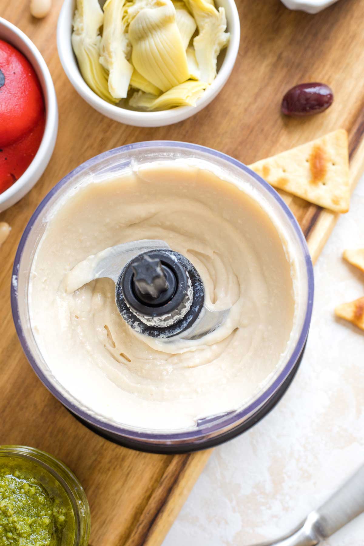 Overhead of base layer smoothly blended in mini food processor with other ingredients around it on cutting board.